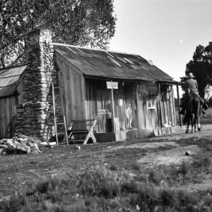 Black and white photograph of a small wooden house with a stone chimney and a man on horseback in front, a white horse next to him and trees in the distance.