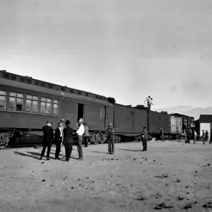 Black and white photograph of a group of men talking next to a passenger train with a white building in the background and mountains in the distance.