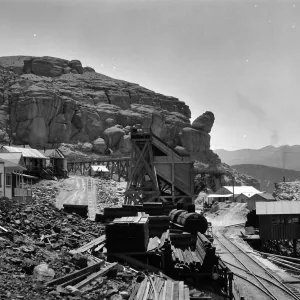 Black and white photograph of train tracks and wooden buildings beside a rocky hill with hills in the distance.