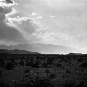Black and white photograph of a desert landscape with rays of sunlight coming through clouds and mountains in the distance.