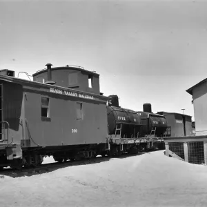 Black and white photograph of a train station, with a wooden building and three black train cars visible. The first car says Death Valley Railroad.