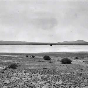 Black and white photograph of a black car on a wide white salt flat with hills in the distance.