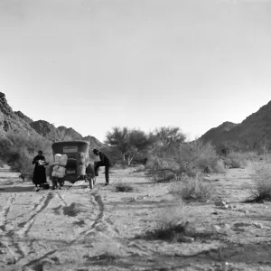 Black and white photograph of a black car and a man and woman in a desert landscape with scrub and low hills around them. The man has his foot propped on the car and the woman is looking at something in her hands.