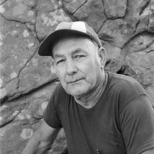 Black and white photograph of a man in a short sleeved shirt and baseball cap looking at the camera against a rock wall background.
