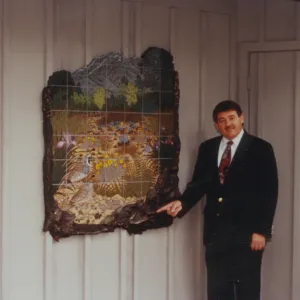 A man in a suit and red tie points at an irregularly shaped mosaic hanging on a white wood paneled wall. The mosaic depicts two California quails in a meadow of long, yellow and dull green grasses and yellow, pink, and pale blue flowers, in front of a background of trees and mountains. A grid pattern is overlaid the mosaic image, and the edges of the piece are deep brown wood carved in the shapes of leaves. 