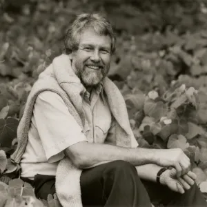 A black and white photo of a bearded man wearing a light-colored shirt and dark slacks with a light-colored sweater over his shoulders. He sits in a field of vines with many heart-shaped leaves. 