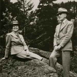 A black and white photo of a man and woman atop a rocky outcropping in front of a forest of evergreen trees. Both wear suits and trilby hats, the woman sits on the outcropping, smiling at the camera, and the man stands to the side looking out toward the distance.