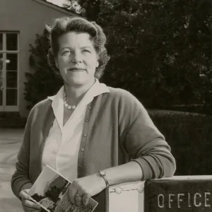 A black and white photo of a woman in a blouse posing in front of a building with neatly trimmed hedges. She holds a booklet depicting a field in front of a forest and mountain and one of her arms rests upon a wooden fence gate with a sign reading "Office" cutoff at the edge of the image.