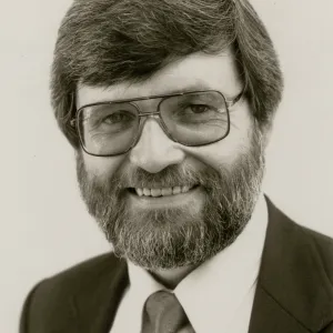 A black and white close-up of a bearded man in a suit and squarish glasses in front of a blank background. 
