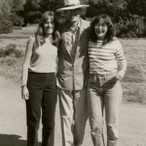A black and white photo of a smiling man wearing a suit and large, wide-brimmed hat and two smiling women in causal dress in a rocky field in front of a copse of trees.