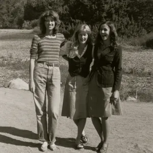 A black and white photo of three jovial women with their arms around each other in a rocky field in front of a copse of trees. 