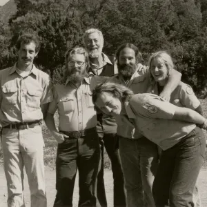 A black and white photo of six smiling people in a rocky field in front of a copse of trees. One man leans in, bent double from the side, in front of the rest of the group.