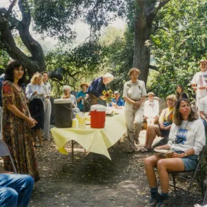 Group of people sitting and standing around table of food in woods looking to the right. 