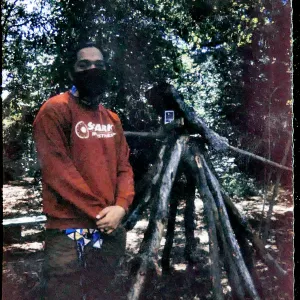 Colored photo of a person wearing medical mask standing next to construction of large sticks in woods. 