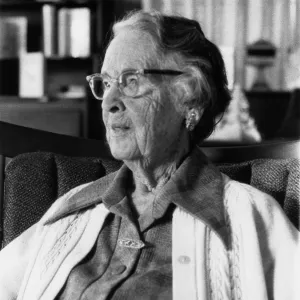 Black and white photograph of woman sitting in chair looking out in distance wearing button up cardigan and glasses.
