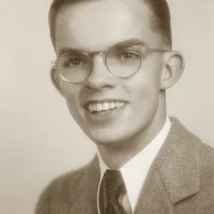 Black and white photograph of young male posing in suit and tie with glasses. 