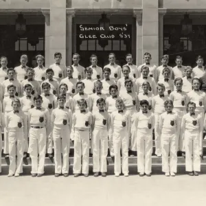 Black and white photograph shows four rows of high school boys in white uniforms standing in front of building with poster. 