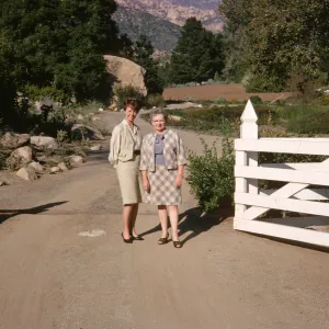 Two women, one older and one younger, in pale colored suits stand, smiling on a gravel road in an open white fence gate. The road behind them is lined with rocks and trees, with mountains and a large boulder visible in the background.