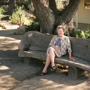 An older woman in a checkered, pale suit smiles while sitting on a large wooden bench at the edge of a stone and gravel path. The bench is made of a large, roughly cut log and sits beneath a tree with a triple trunk. 