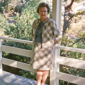An older woman in a pale, checkered suit stands on a shady porch against a white, wooden railing. 