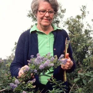 An older woman wearing glasses and a green shirt and navy sweater stands in waist high vegetation. She is examining a thin branch laden with clusters of lavender colored flowers and has a clipboard tucked under her left arm. 