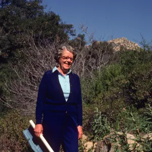 An older woman dressed in royal blue smiles in front of a rocky hill coated in short vegetation and bare-branched bushes. A rolled blueprint is held in her right hand. 