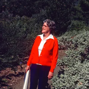 An older woman in a bright orange-red sweater stands on a path flanked on both sides by tall bushes, smiling into the distance. She holds a rolled set of blueprints in her right hand. 