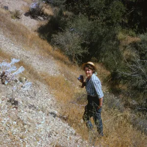 A woman holding a camera wearing a blue and white striped shirt and straw hat stands on a steep, rocky incline striped with brown grasses.  
