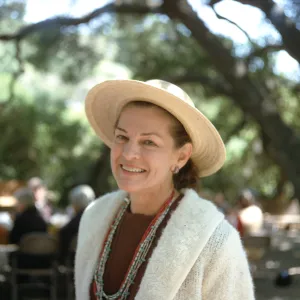 A woman in a straw hat, white cardigan, and numerous beaded necklaces smiles at the camera. In the background, several people sit at an outdoor table beneath a large double-trucked tree.