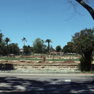 A park consisting of grassy areas and beds of soil dotted with large stones separated by paths viewed from across a paved street. 