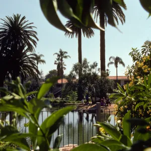 On the far side of a large pond, several people are enjoying a park, walking along paths or sitting on a bench in the shade of a tree at the edge of the pond. 